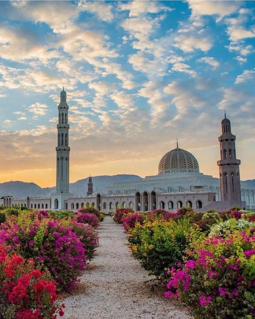 The Sultan Qaboos Grand Mosque at dawn, its tall main minaret and tiled dome pale against a cloud-streaked sky, with a gravel path between banks of pink and red bougainvillea leading towards it