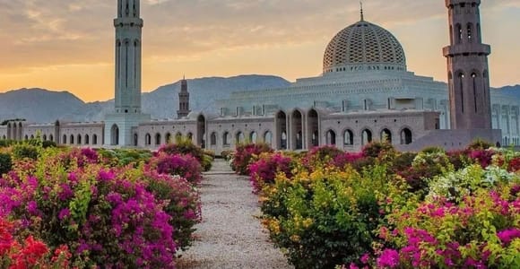 The Sultan Qaboos Grand Mosque at dawn, its tall main minaret and tiled dome pale against a cloud-streaked sky, with a gravel path between banks of pink and red bougainvillea leading towards it