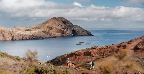 Three walkers on a red-brown path above a bay, with a rocky headland with a natural arch, a sailing boat and fish-farm pens on the water