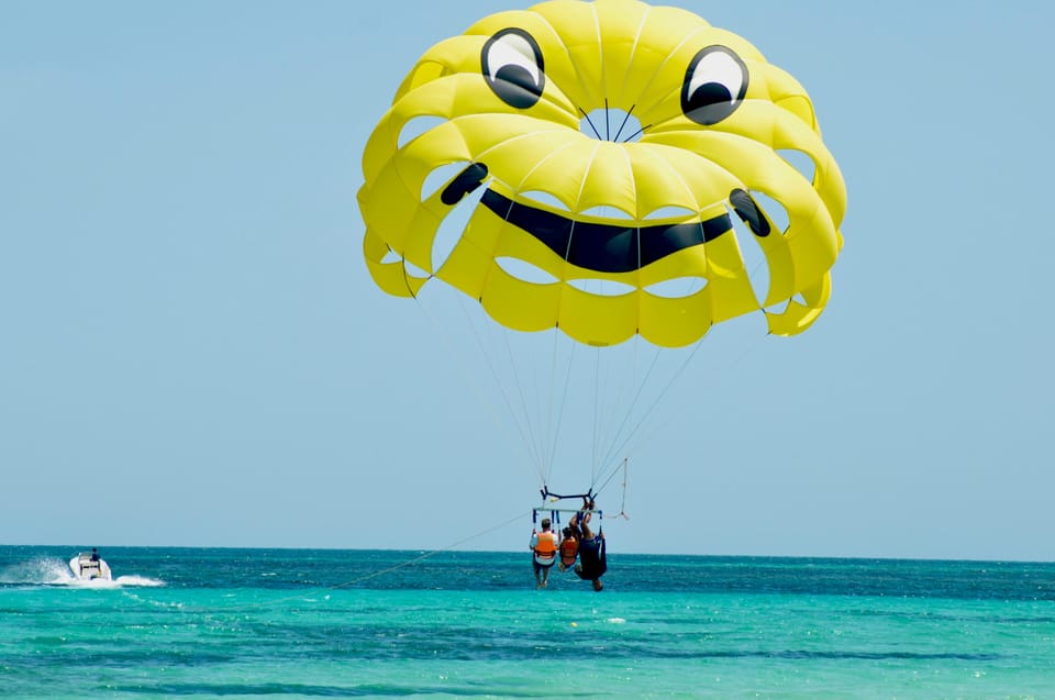 A yellow parachute with a smiling face above a turquoise sea, three people in orange life jackets hanging below it and a small boat in the distance