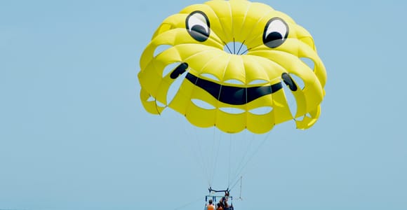 A yellow parachute with a smiling face above a turquoise sea, three people in orange life jackets hanging below it and a small boat in the distance
