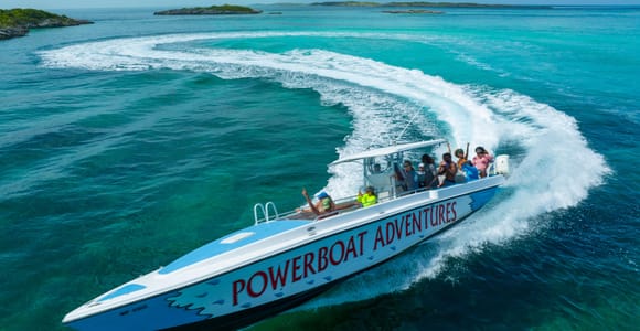 An aerial view of a white speedboat lettered Powerboat Adventures carving a wide turn through turquoise shallows, with low green cays on the horizon