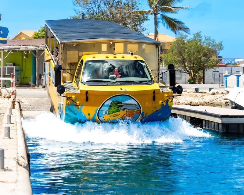 A yellow and blue amphibious bus driving off a concrete slipway into calm water, throwing up a wall of white spray, with pastel houses and palms behind it