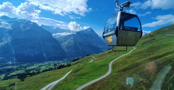 A grey gondola cabin above green alpine meadows with a snow-capped Eiger ridge in the background