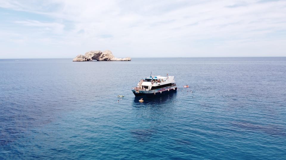 White excursion boat anchored in open blue water off a rocky islet, swimmers floating around it