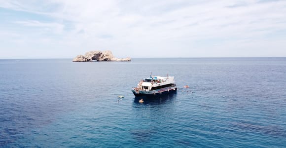 White excursion boat anchored in open blue water off a rocky islet, swimmers floating around it