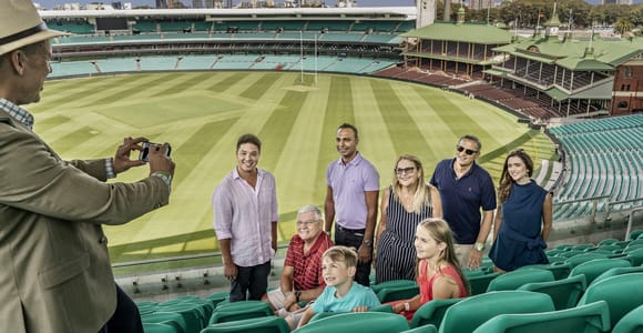 A guide photographing a group of visitors seated in empty green stands above a striped cricket outfield, with a heritage brick pavilion beyond