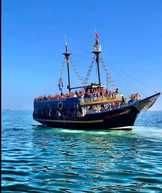 A dark wooden pirate-style ship crowded with passengers on bright turquoise water under a clear blue sky