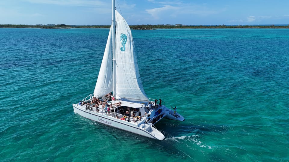A white sailing catamaran with its mainsail up crossing calm blue-green water under a clear sky, seen from another boat