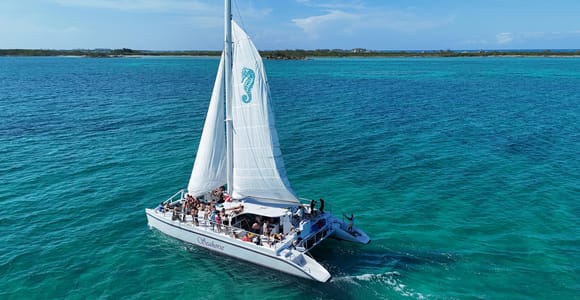A white sailing catamaran with its mainsail up crossing calm blue-green water under a clear sky, seen from another boat