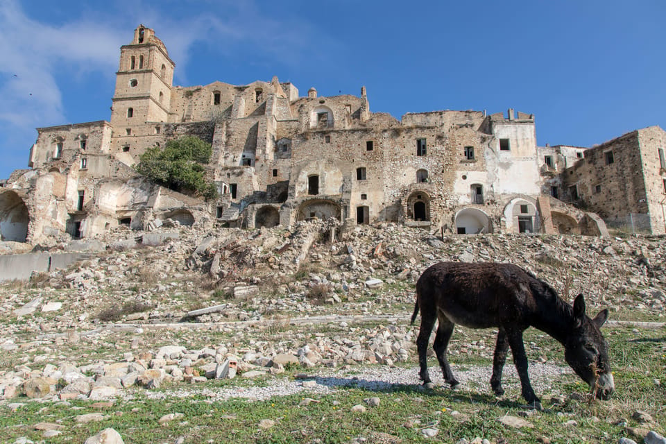 Abandoned stone buildings of Craco above a rubble slope, with a donkey grazing in front
