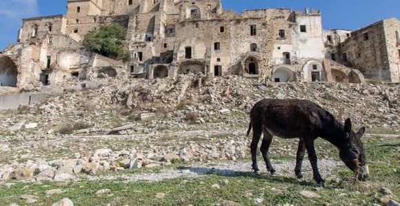 Abandoned stone buildings of Craco above a rubble slope, with a donkey grazing in front