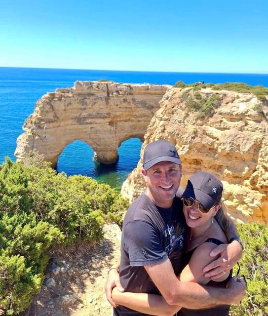 A couple in caps hug on a clifftop above the sea, with a heart-shaped rock arch and blue water behind them