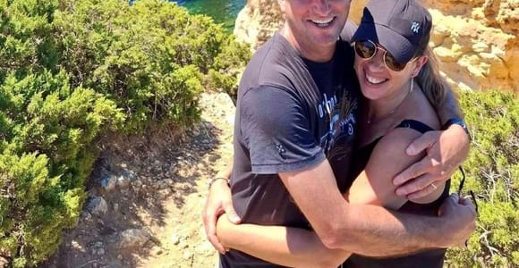 A couple in caps hug on a clifftop above the sea, with a heart-shaped rock arch and blue water behind them