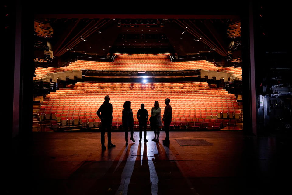 Five people in silhouette standing on a darkened stage, looking out at rows of empty orange seats lit from above