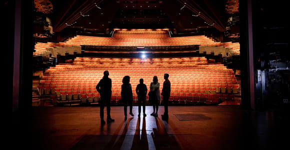 Five people in silhouette standing on a darkened stage, looking out at rows of empty orange seats lit from above