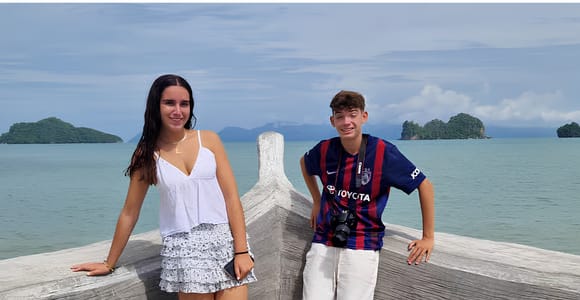 Two teenagers standing at the pale stone plinth of the eagle monument at Kuah, small forested islands scattered across the bay behind them