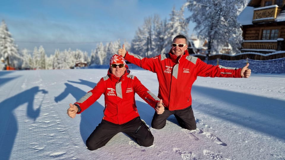 Two ski instructors in red jackets kneeling on a groomed snowy slope, giving thumbs up