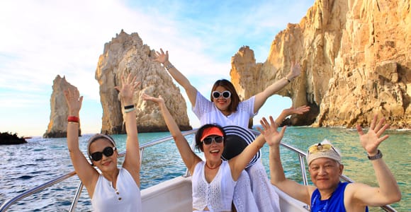 Four passengers with their arms raised on an open boat in front of the Land's End arch, turquoise water around them