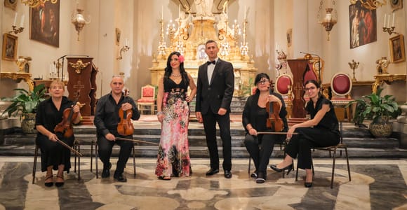 A string ensemble and two singers in formal dress posed on the steps in front of a gilded altar in a church