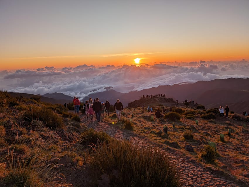 Walkers on a stony path along a mountain ridge towards a lookout, with the low sun above a sea of clouds