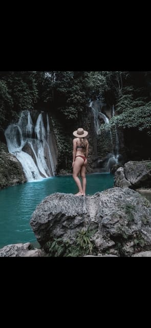 A woman in a straw hat standing on a boulder above a turquoise plunge pool, with a wide fan of waterfall strands and dense dark jungle behind her