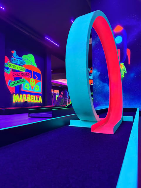 Glowing neon minigolf course with a red and turquoise loop and a painted Marbella mural on the wall