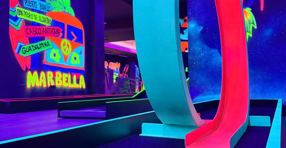Glowing neon minigolf course with a red and turquoise loop and a painted Marbella mural on the wall