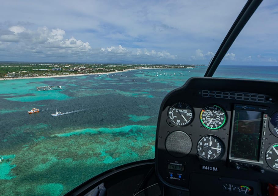 View from a helicopter cockpit over turquoise reef shallows, moored boats and a line of hotels on the horizon, instrument dials to the right