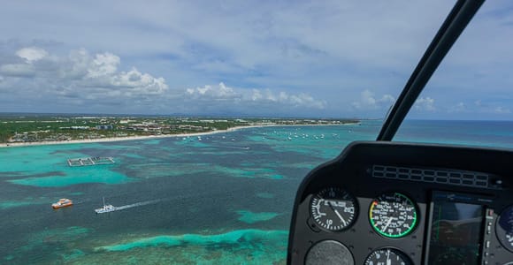 View from a helicopter cockpit over turquoise reef shallows, moored boats and a line of hotels on the horizon, instrument dials to the right