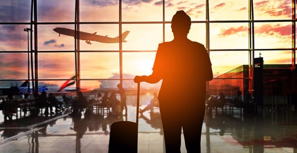 Silhouette d’un voyageur avec une valise à roulettes devant la baie vitrée d’un aéroport au coucher du soleil, un avion décollant derrière
