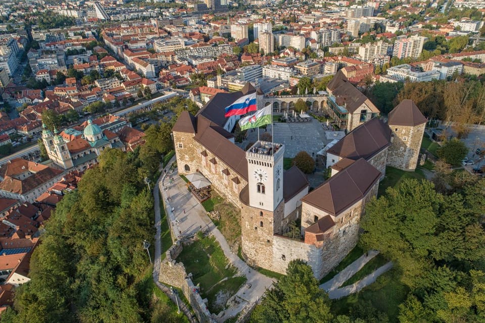 Aerial view of a stone castle with a clock tower and a Slovenian flag above the red rooftops of the old town