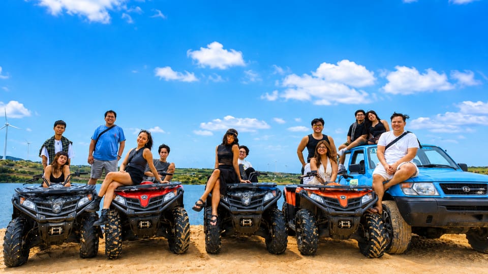 A large group posing on four quad bikes and a blue open-top jeep on sand beside a lake, wind turbines on the far shore