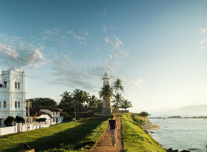 A person walking a grass path along sea-facing ramparts towards a white lighthouse among palms, a white colonial building on the left and the ocean on the right