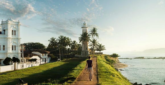A person walking a grass path along sea-facing ramparts towards a white lighthouse among palms, a white colonial building on the left and the ocean on the right
