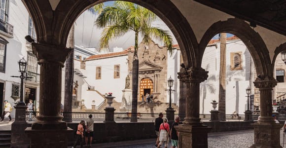 View through stone arches onto a cobbled street, palm trees and a church façade with a flight of steps in Santa Cruz de La Palma