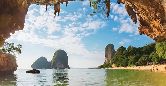 Longtail boats moored off a sandy beach beneath an overhanging limestone headland at golden hour