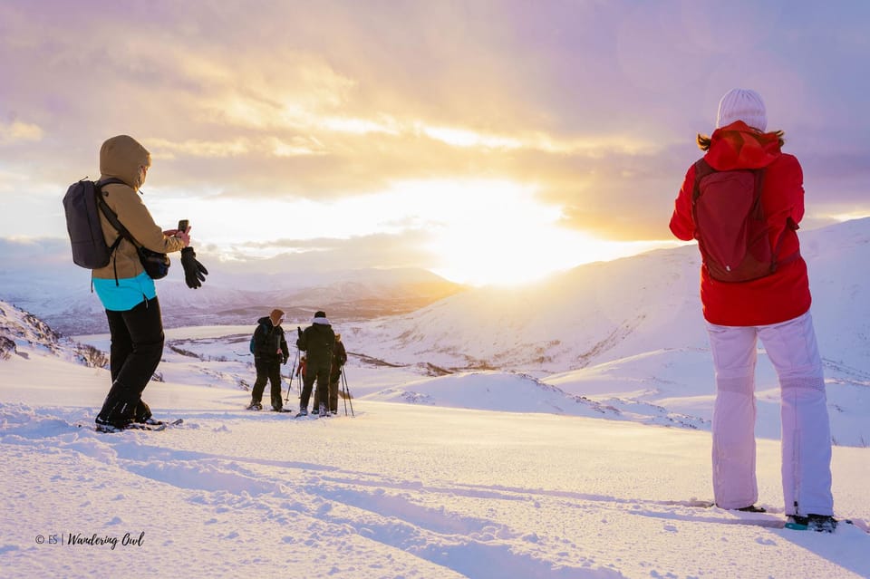 Hikers on a snow-covered ridge photographing a low sun glowing behind clouds over distant mountains