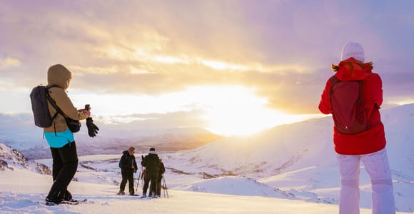 Hikers on a snow-covered ridge photographing a low sun glowing behind clouds over distant mountains