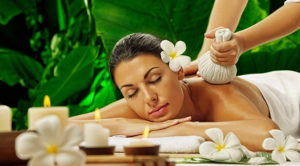 A woman with a white flower in her hair lies on her front with closed eyes while a hand presses a herbal compress to her back, candles and white frangipani flowers in front, green leaves behind