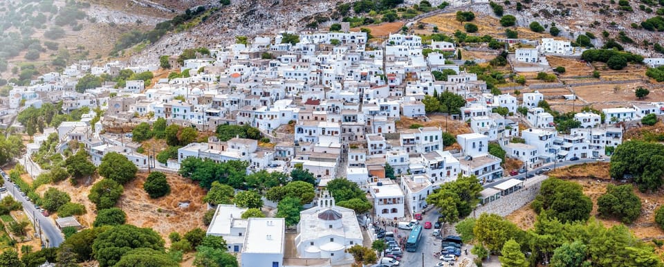 A white village climbing a rocky hillside, with a bus and parked cars at its lower edge