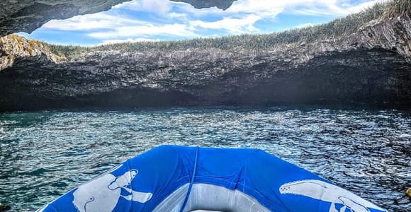 View from the bow of an inflatable boat with a humpback whale painted on its tube, looking out of a rock arch towards grass-topped cliffs
