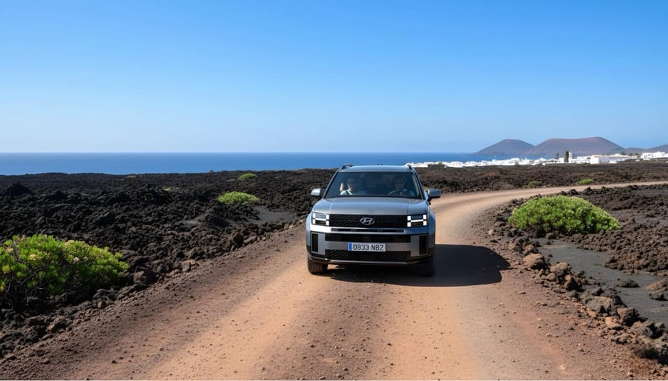 A silver hire car coming towards the camera on a graded dirt track between black lava fields, with the sea on the left and white houses below a volcano behind