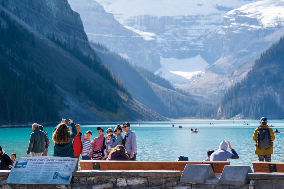 Visitors on the Lake Louise shore looking over turquoise water towards a glaciated valley, with canoes in the distance