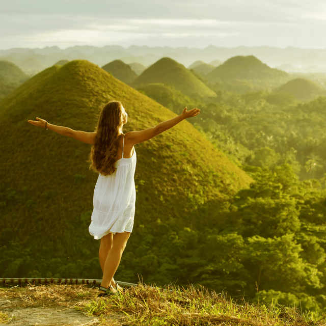 A woman in a white dress standing on a grassy ridge with her arms spread wide, rows of green hills fading into haze behind her