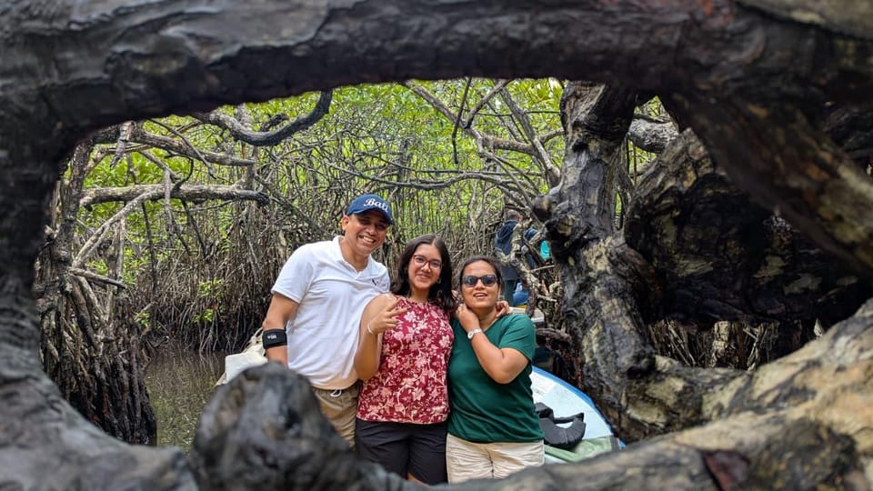 A family of three standing inside the hollow of a huge mangrove root, their boat moored in the green channel behind them