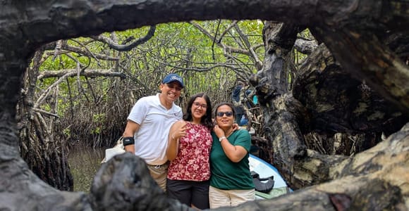 A family of three standing inside the hollow of a huge mangrove root, their boat moored in the green channel behind them