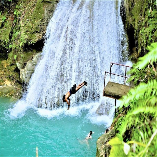 A person diving head-first from a metal platform into a turquoise pool at the foot of a wide waterfall, ferns framing the shot