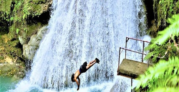 A person diving head-first from a metal platform into a turquoise pool at the foot of a wide waterfall, ferns framing the shot