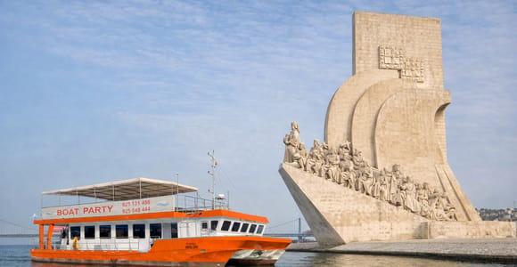 An orange excursion catamaran with BOAT PARTY lettered on the side panel below its white sun roof, passing a pale stone monument shaped like a ship's prow, the Ponte 25 de Abril faint on the horizon to the left